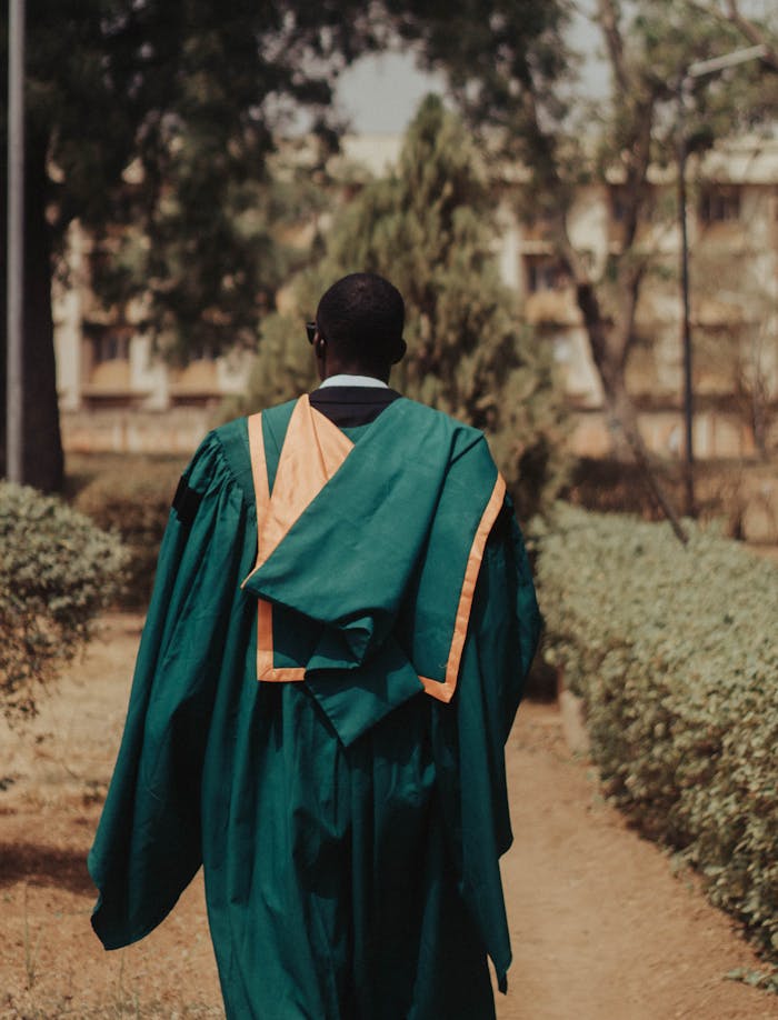 services-01 A graduate in green and orange gown walking outdoors, from behind.